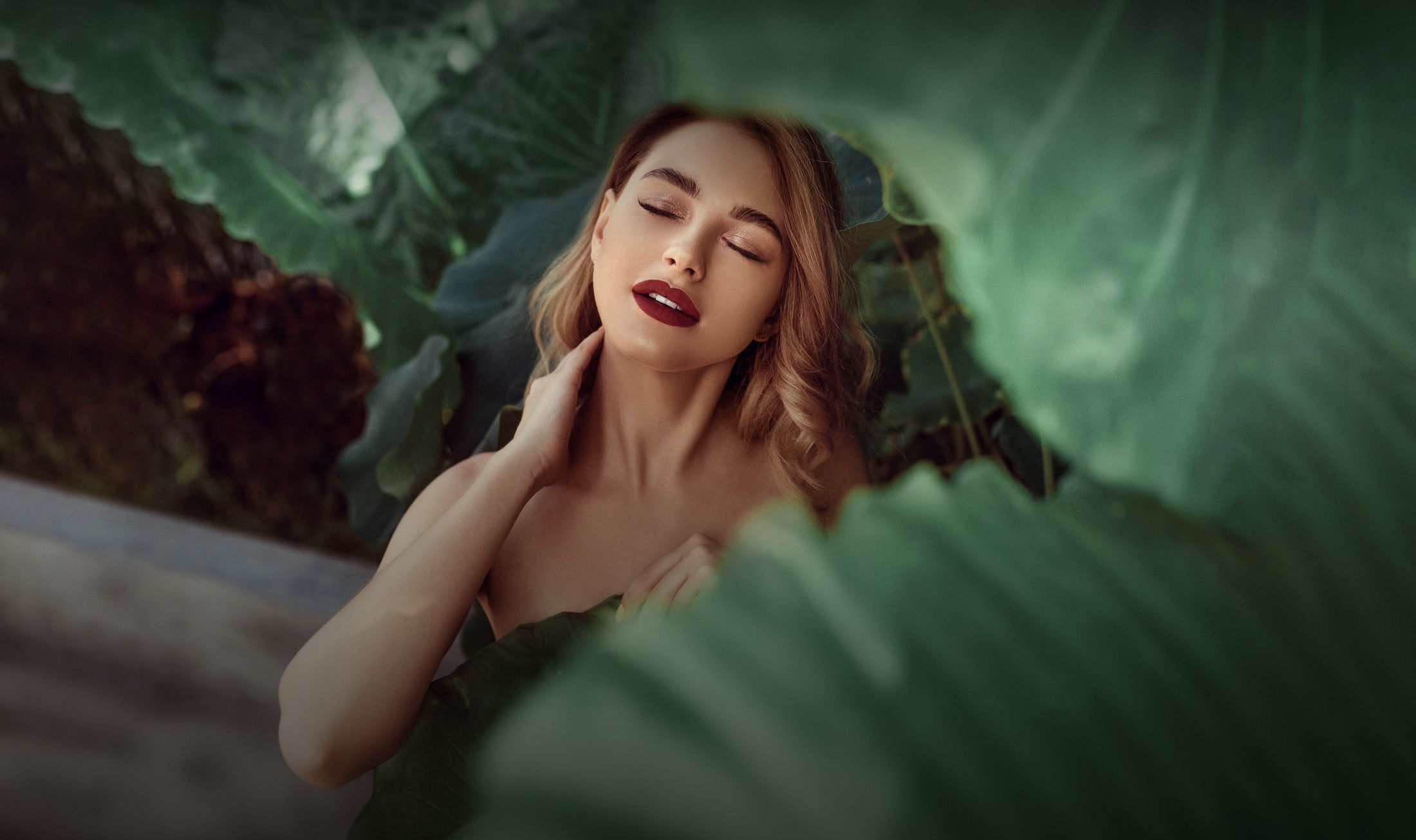 Woman surrounded by lush green leaves, serene expression.